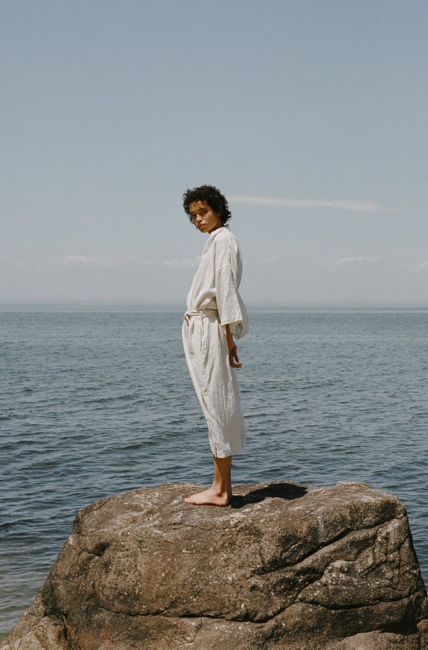 Female model standing on rock wearing the 02 robe - oatmeal by Deiji Studios against ocean horizon