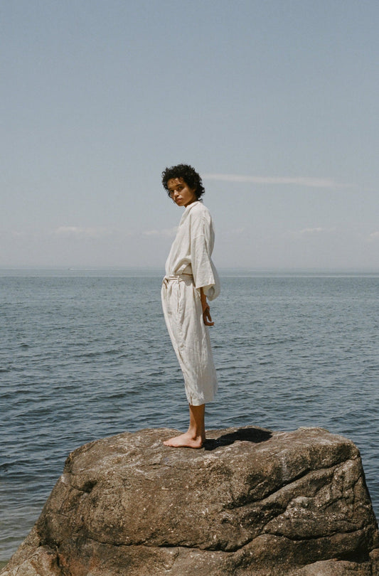 Female model standing on rock wearing the 02 robe - oatmeal by Deiji Studios against ocean horizon