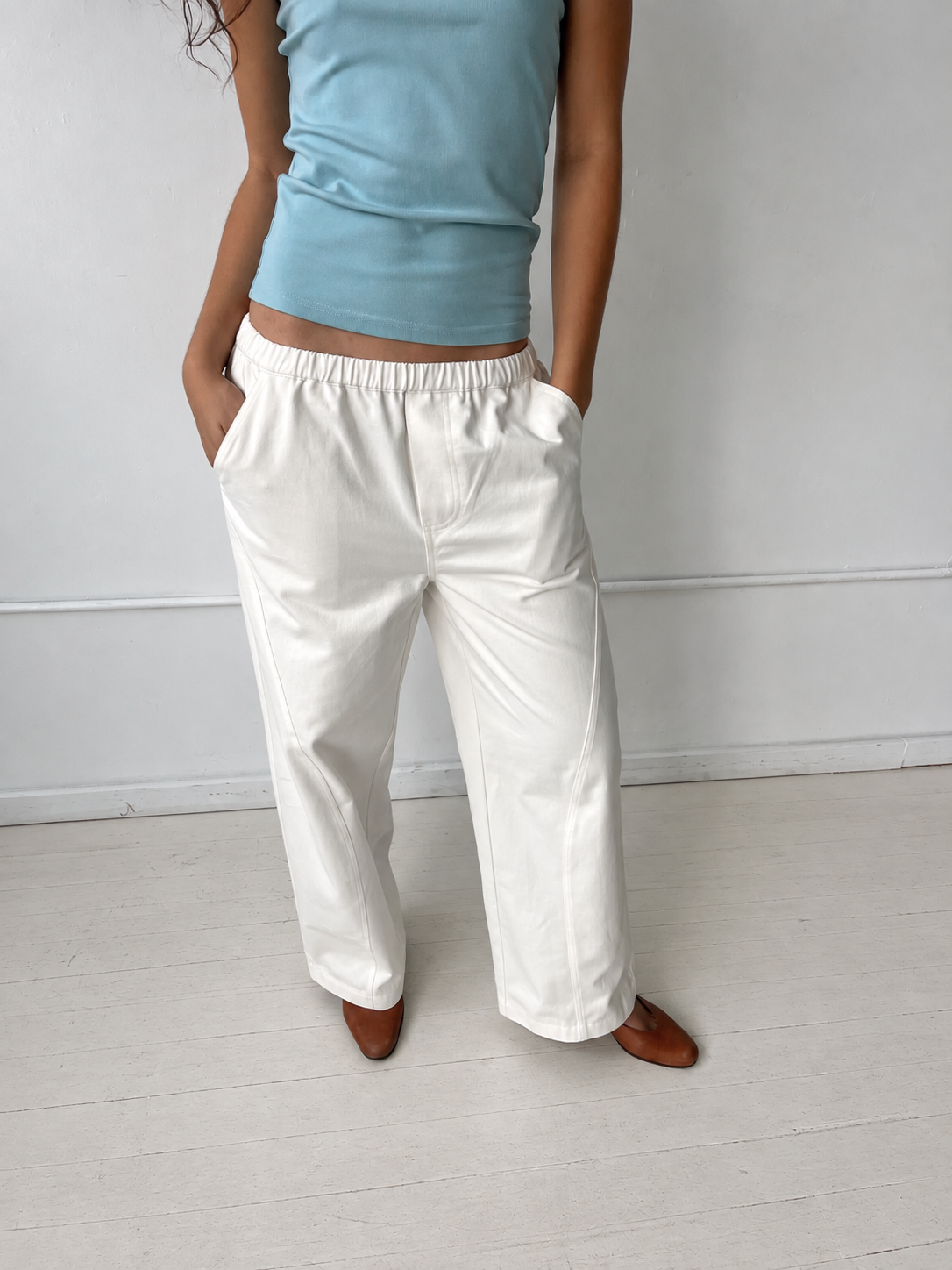 Cropped to the lower body, a woman stands against a white wall with hands in the pockets of the Curve Pant - White by Deiji Studios, a softly structured pant with a relaxed fit and side pockets, styled with the Scallop Tank in Sky.