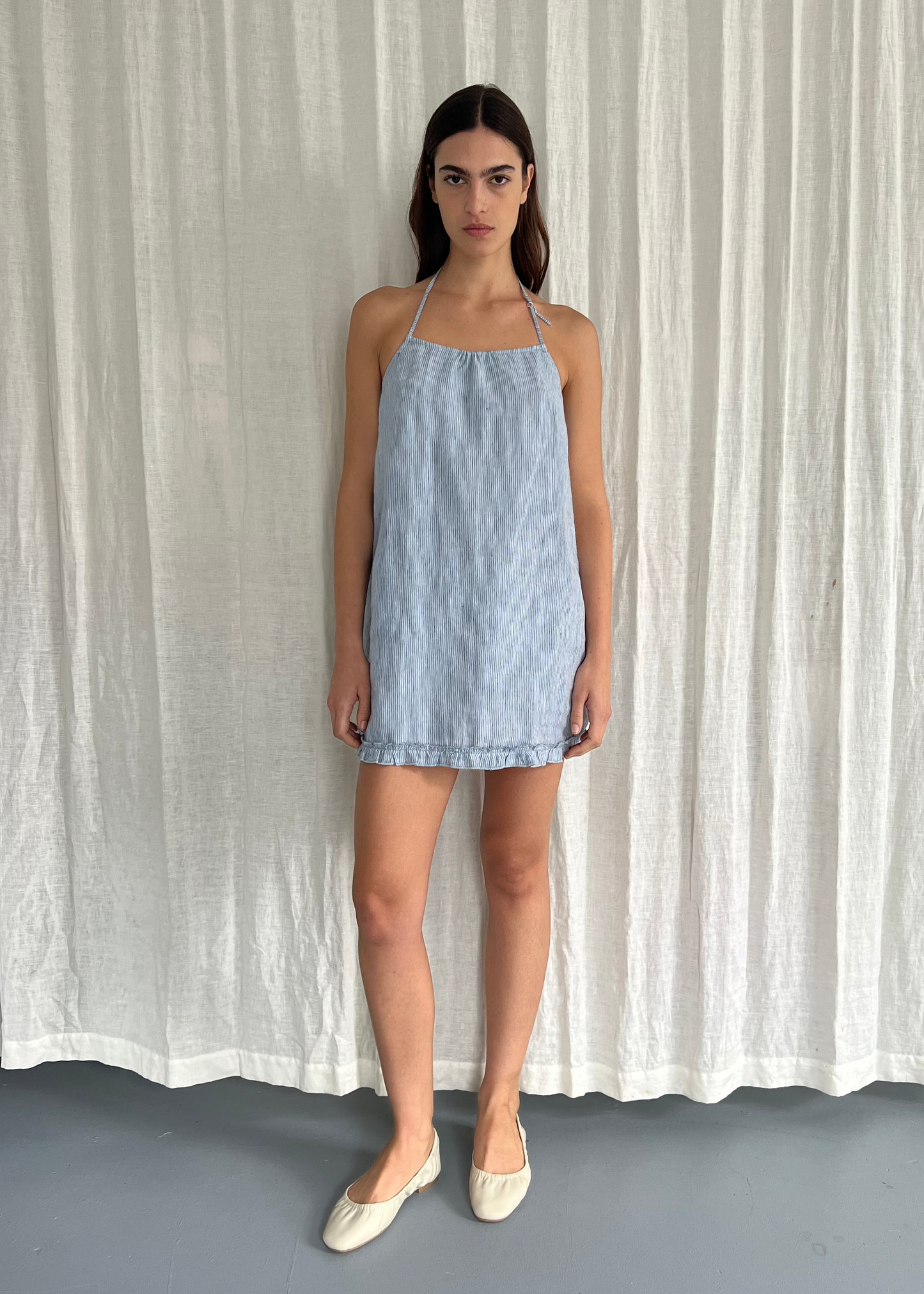 A woman stands before white curtains, wearing the Flounce Halter Dress - Blue Stripe by Deiji Studios, crafted from organic French linen. She faces forward neutrally, arms relaxed by her sides, and wears beige flats.