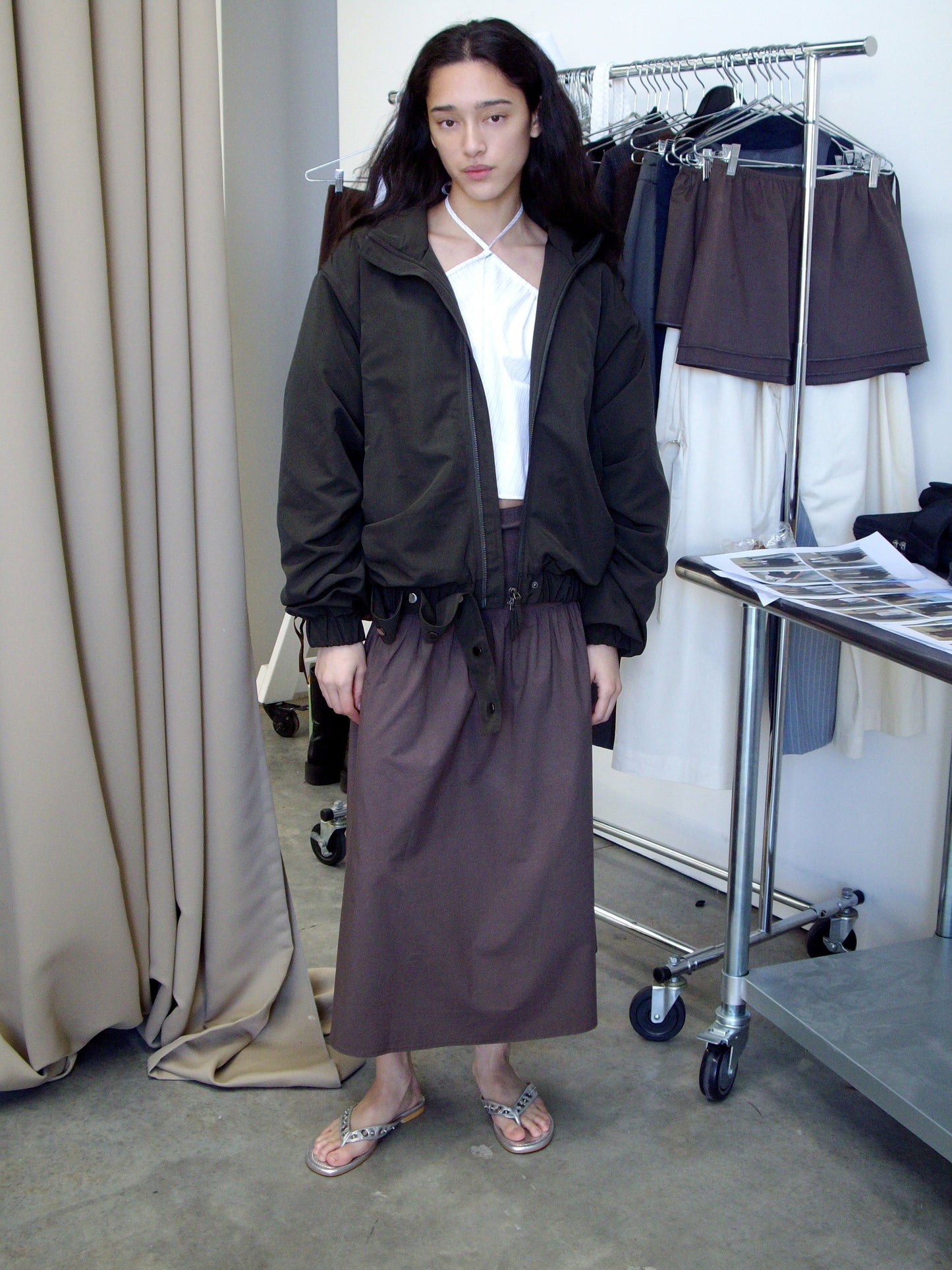 A person with long hair wears a Deiji Studios dark jacket, a white top, the Button Skirt - Chocolate, and flip-flops. They stand indoors by clothing racks and beige curtains, with folded garments and papers on a metal table.