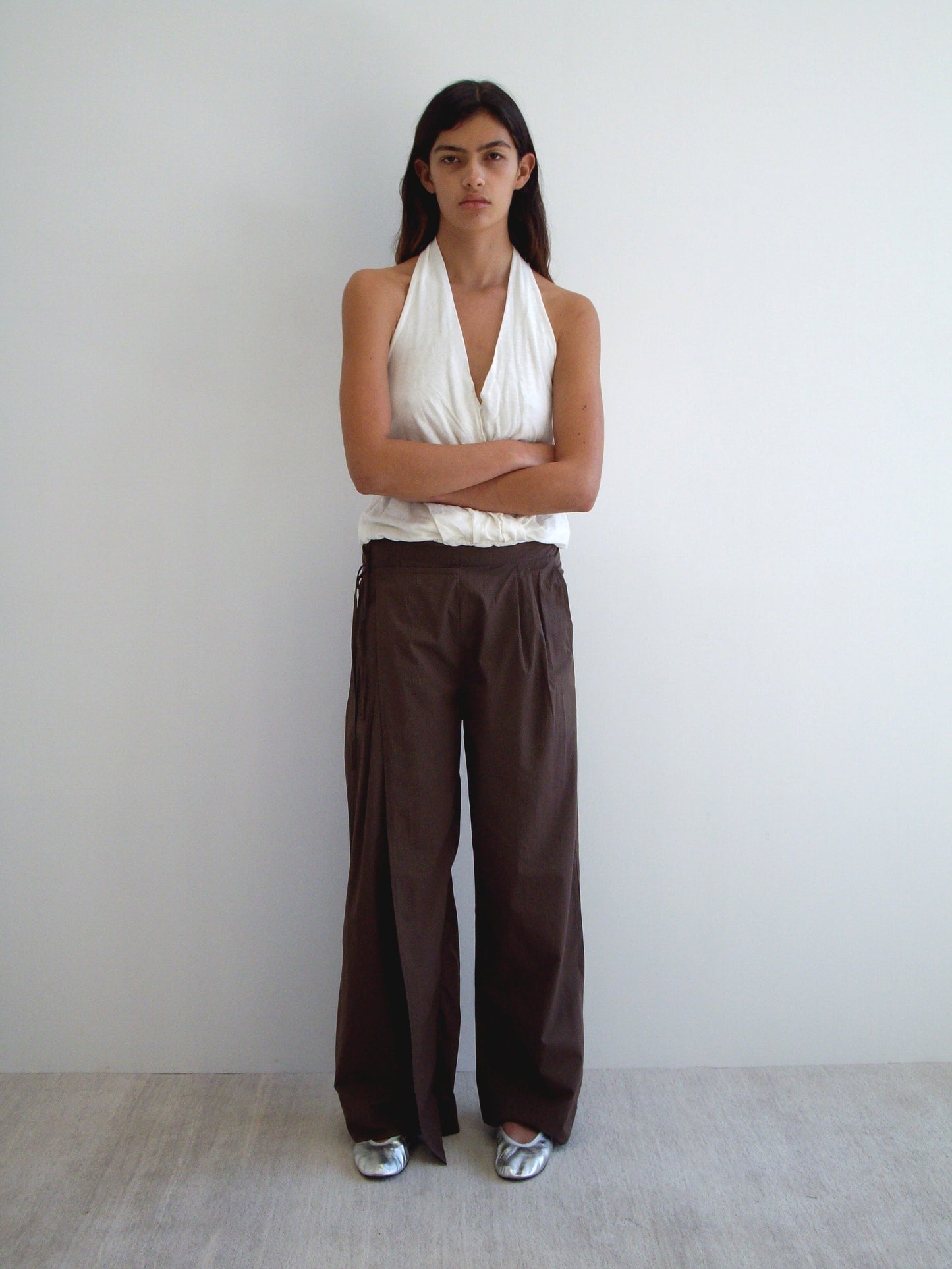 A person stands against a plain white wall with arms crossed, wearing a sleeveless white halter top, Deiji Studios Double Layer Pant - Carob Check in loose brown organic cotton, and silver shoes. They have long dark hair and a neutral expression.