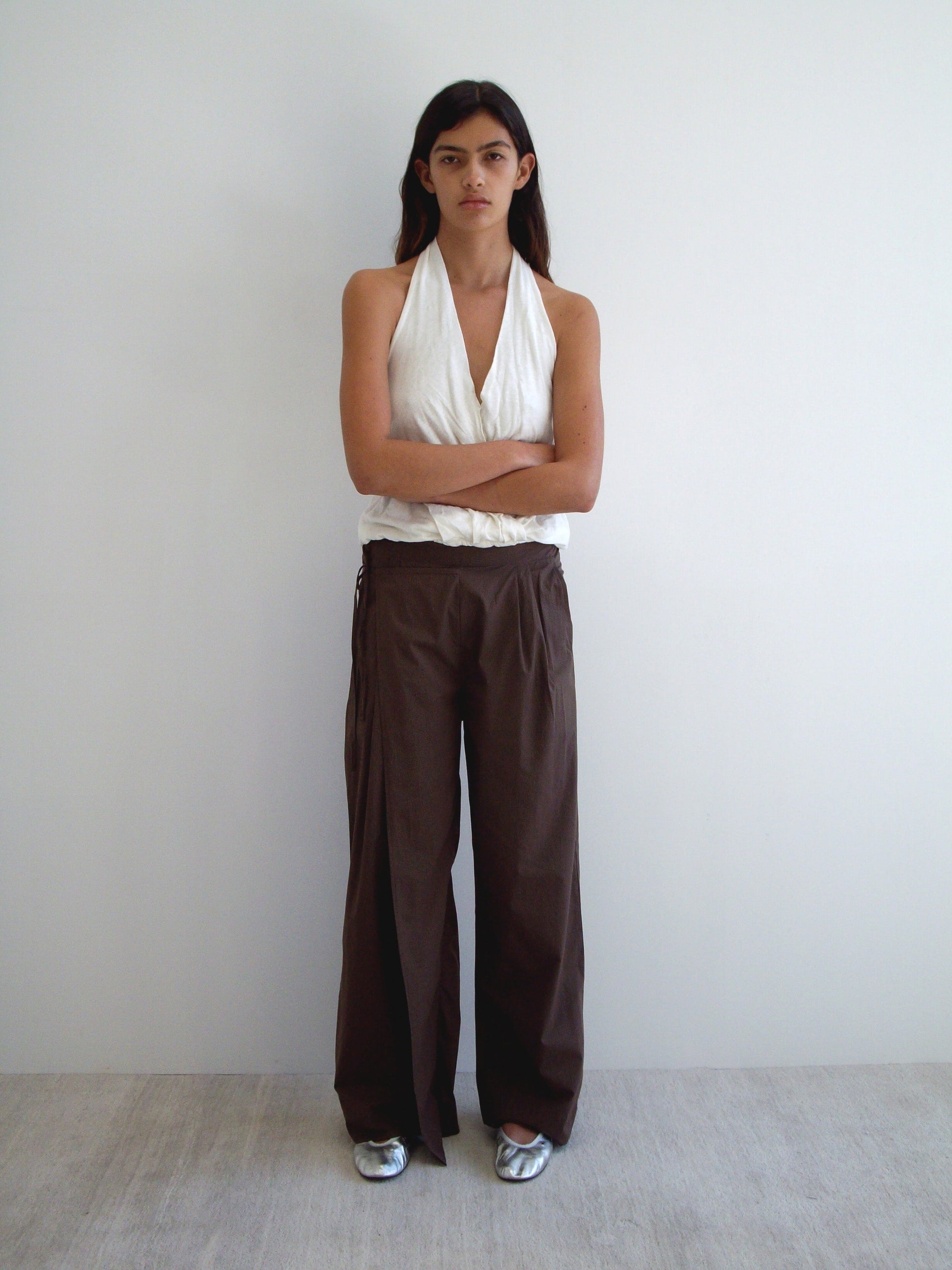 A person stands against a plain white wall with arms crossed, wearing a sleeveless white halter top, Deiji Studios Double Layer Pant - Carob Check in loose brown organic cotton, and silver shoes. They have long dark hair and a neutral expression.