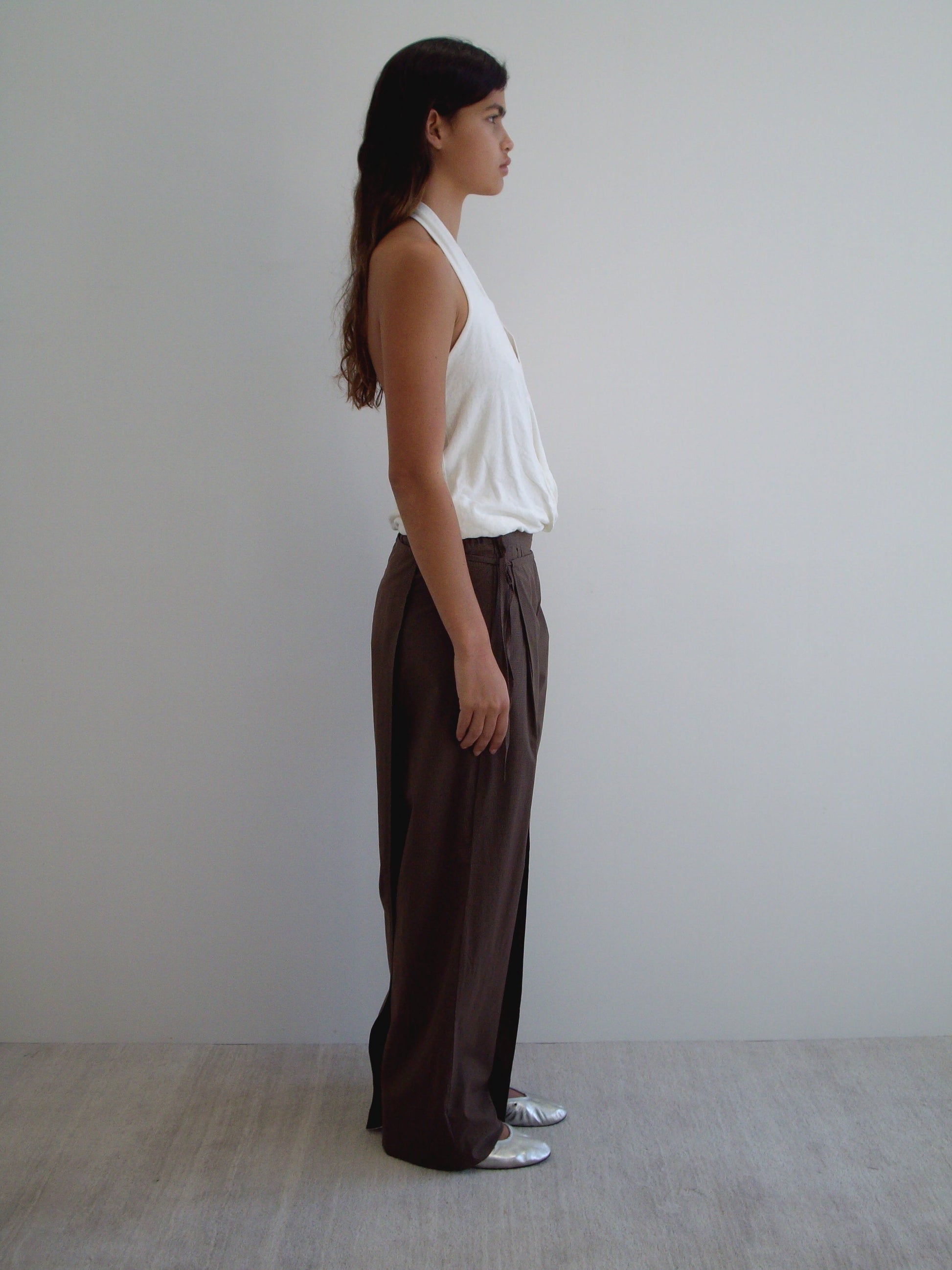 A person with long hair stands in profile against a minimalist background, wearing a sleeveless white top, Double Layer Pant - Carob Check, and silver shoes. The setting features a neutral floor and simple wall.