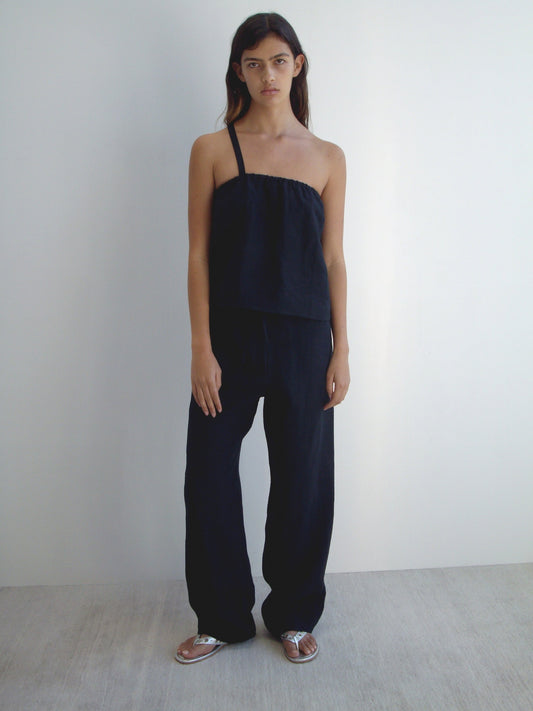A person with long dark hair and a neutral expression stands against a plain white wall, wearing the Button Strap Top - Black by Deiji Studios in organic French linen, paired with loose black pants and silver sandals.