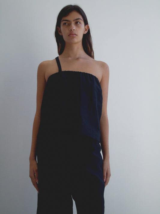 A woman with long dark hair stands against a white background, wearing the Button Strap Top - Black by Deiji Studios and black pants. She has a neutral expression and her arms rest at her sides.