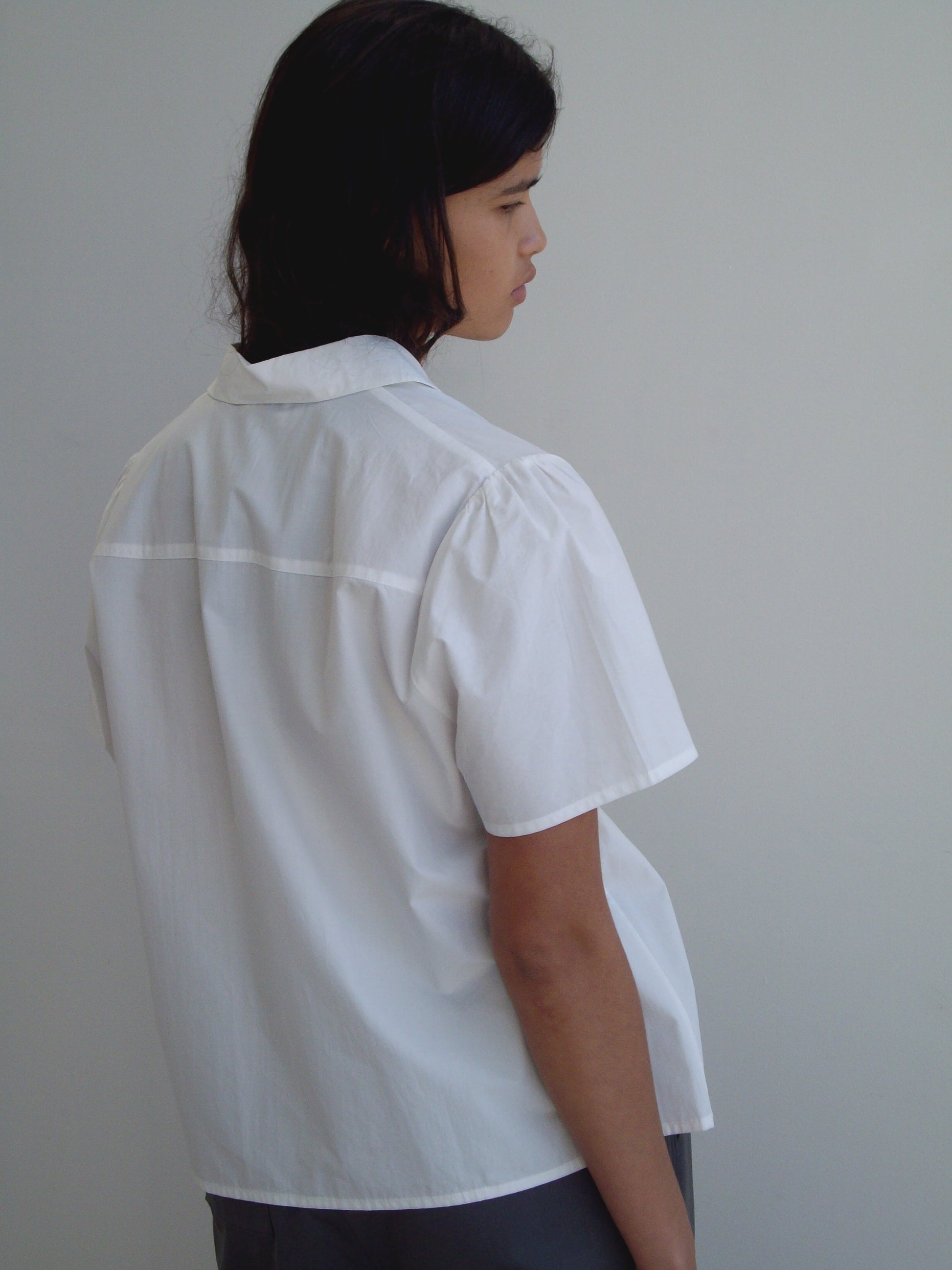 A person with shoulder-length dark hair stands sideways, facing away from the camera, wearing the Deiji Studios Sails Top in white and gray pants against a plain light gray background.