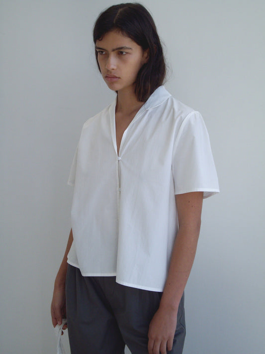 A person with shoulder-length dark hair wears the Sails Top - White from Deiji Studios with gray pants, standing against a plain light wall and looking slightly downward.