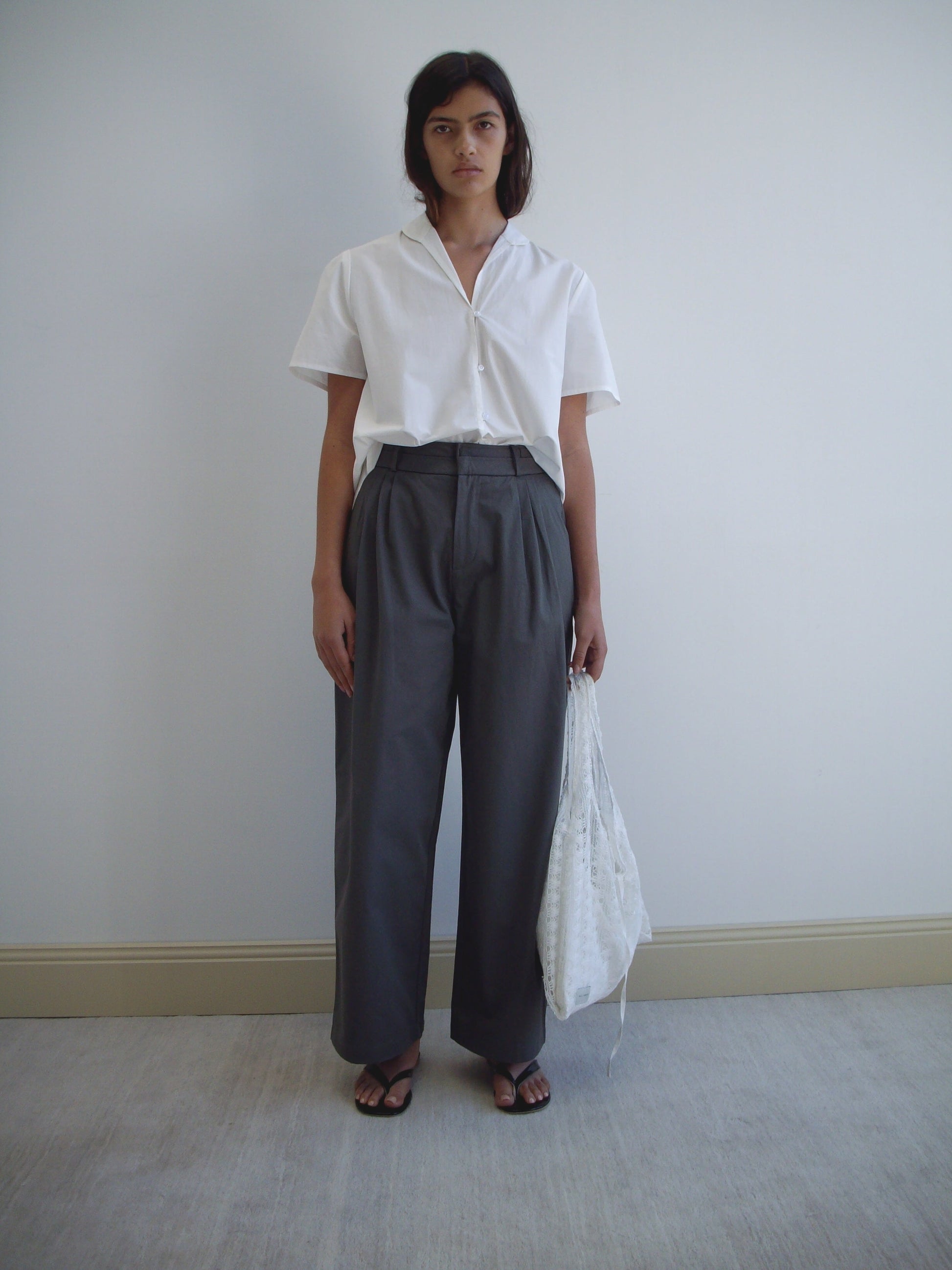 A woman stands against a plain wall wearing a short-sleeved white button-up, Deiji Studios Two Layer Pant - Charcoal, black sandals, and holding a white textured bag at her side.