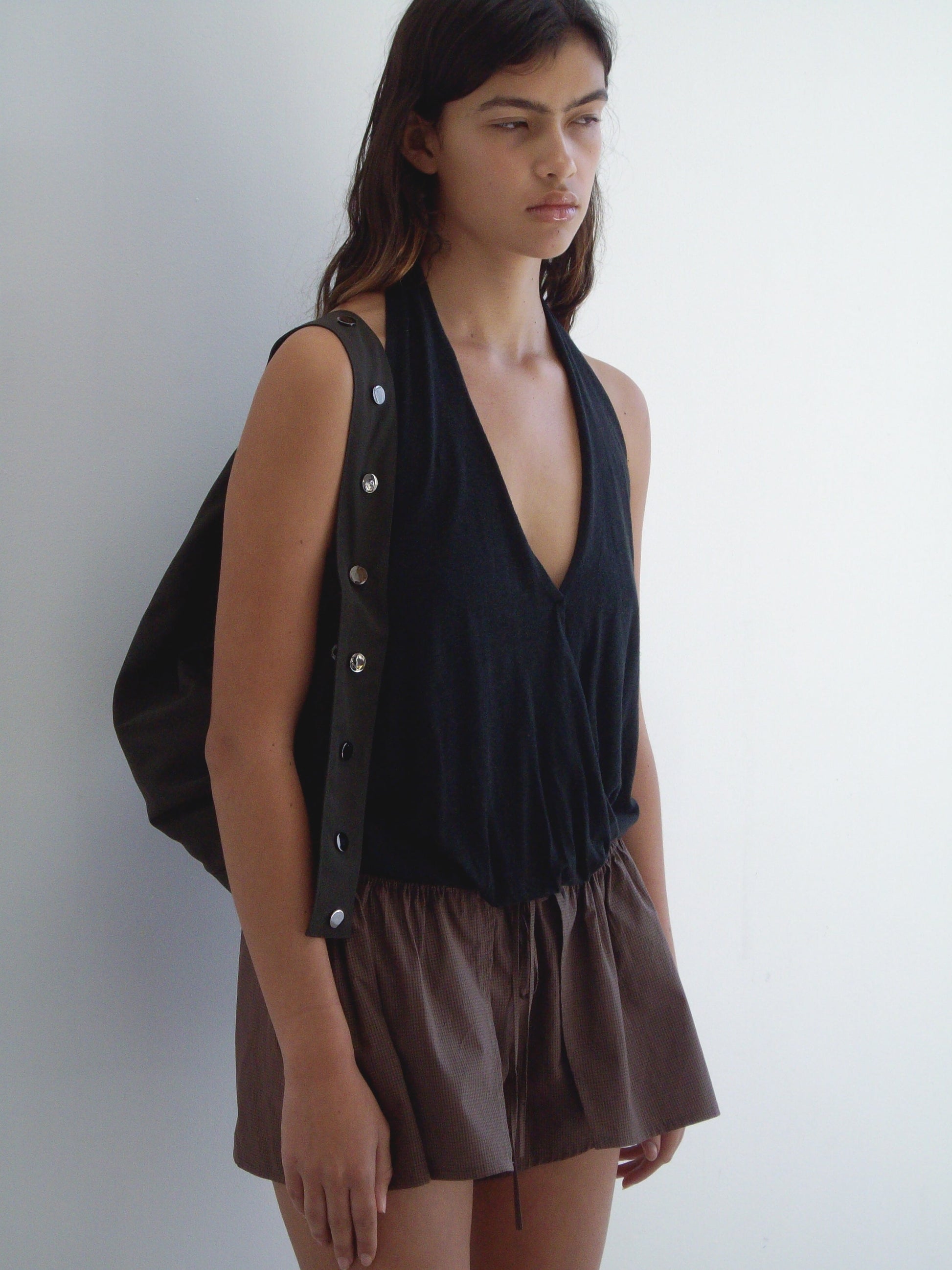 A person with long dark hair stands against a white wall, wearing the sleeveless black Jersey Top with button details on one strap and loose brown shorts, carrying a black bag over one shoulder.