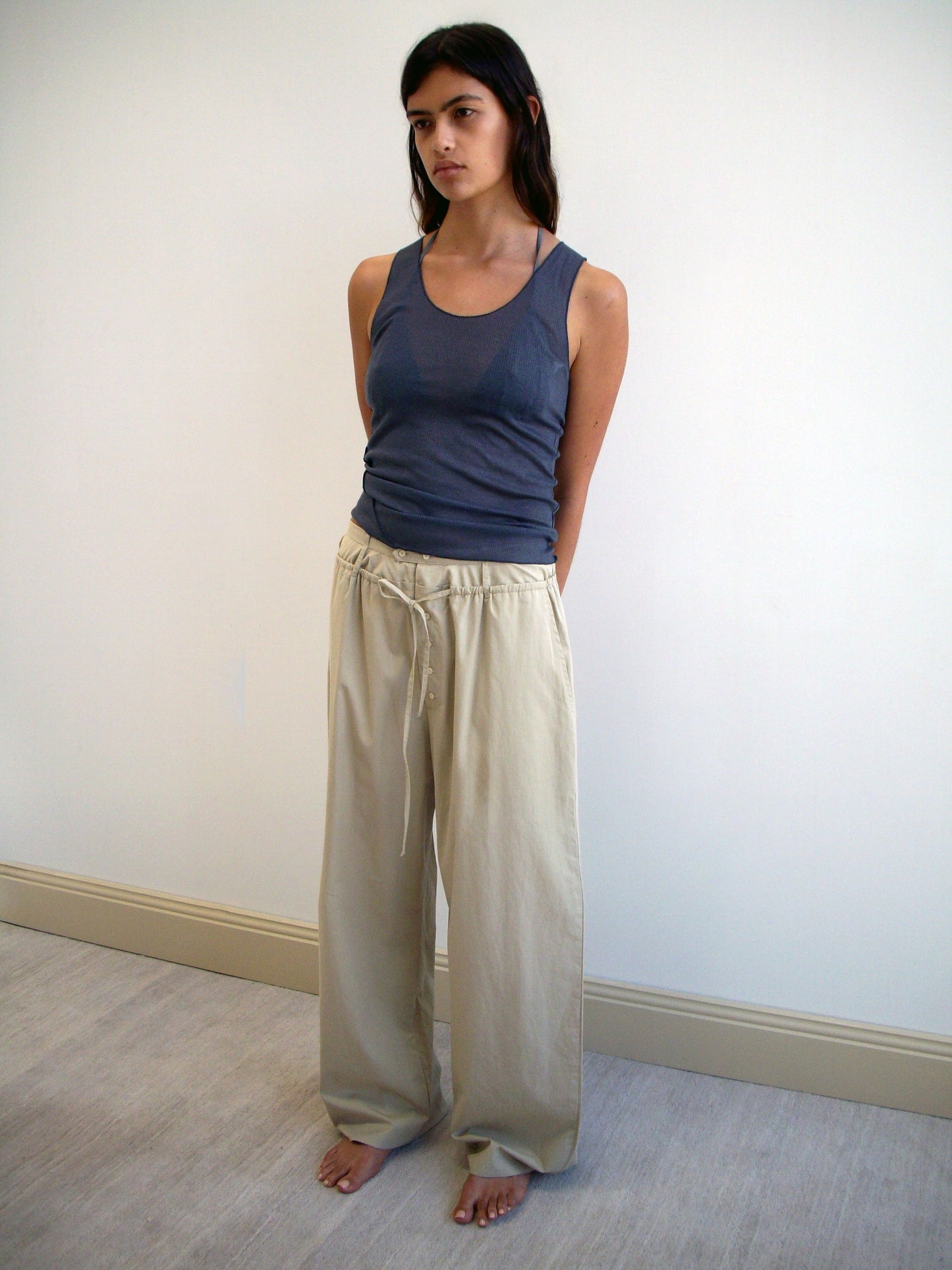 A woman with long dark hair stands barefoot indoors against a plain wall, wearing a blue tank top and loose Two Band Pant - Fawn by Deiji Studios. Her arms are relaxed behind her back.