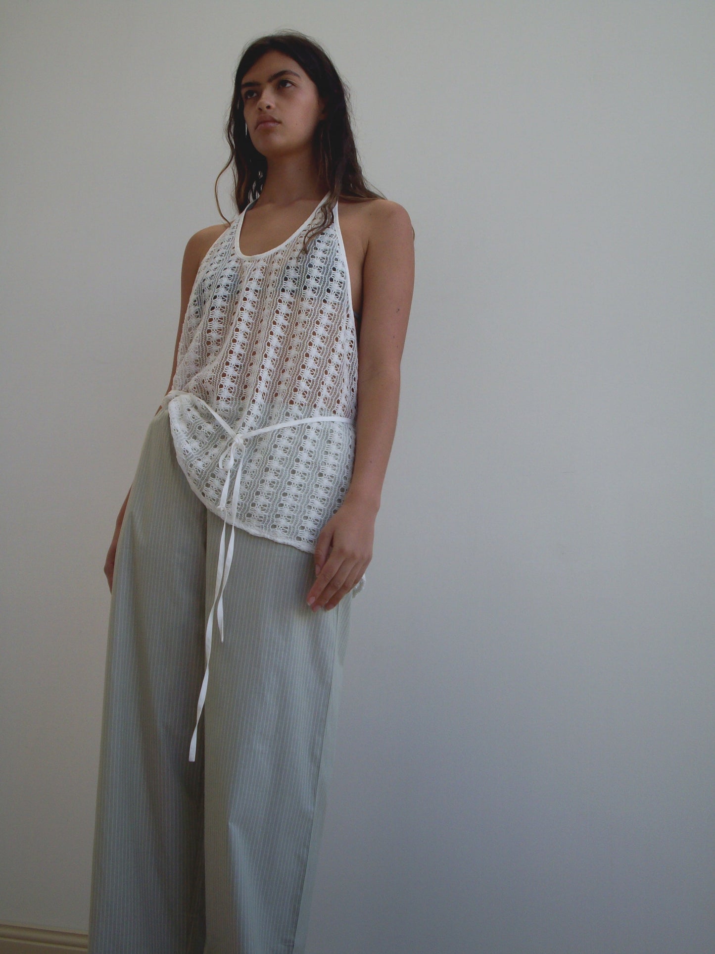 A woman stands against a plain white wall, wearing a loose, sleeveless, sheer Deiji Studios crochet top over a white bralette and Ease Trouser in Pistachio Stripe. She gazes thoughtfully off to the side.