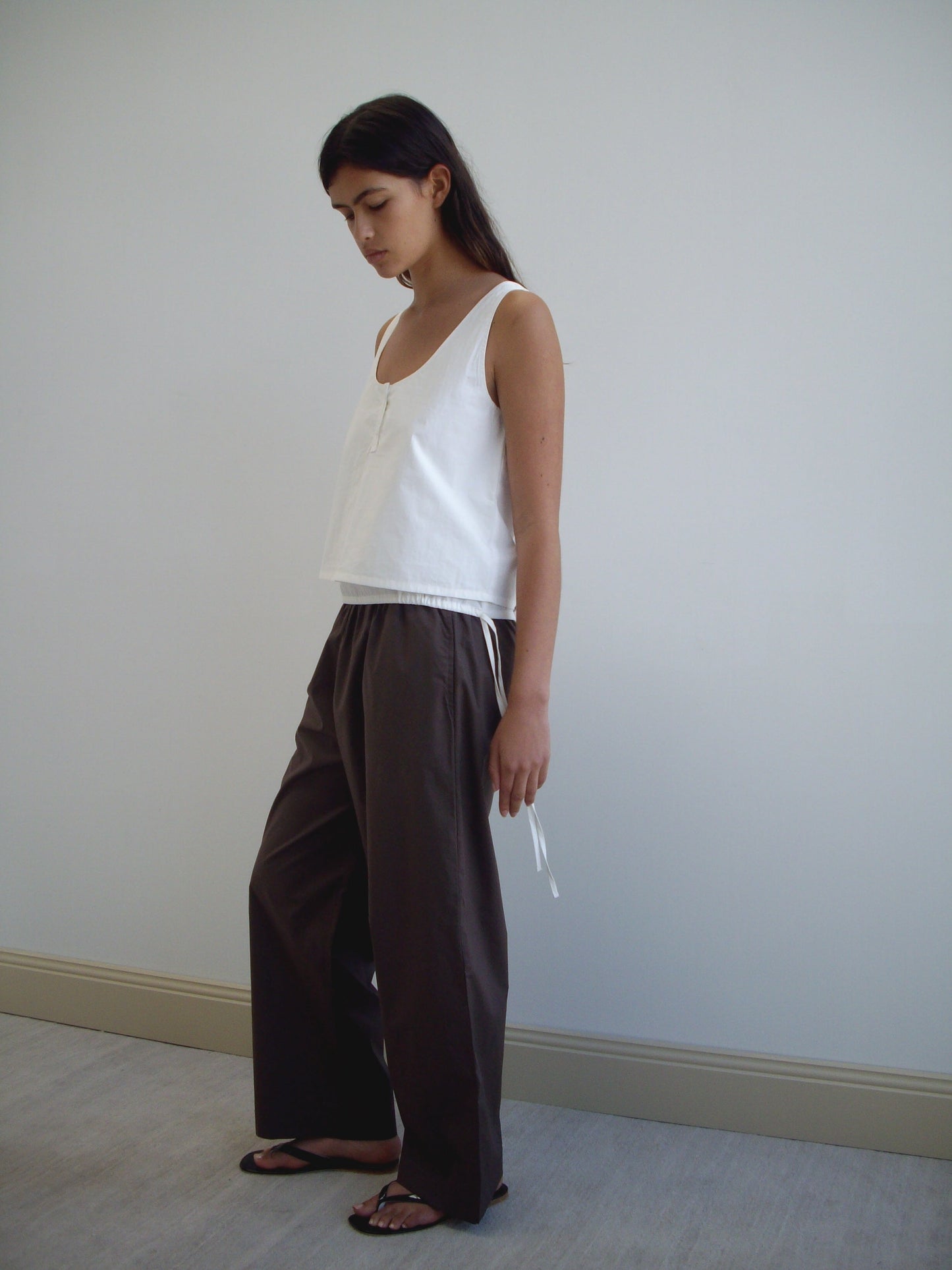 A woman stands indoors against a plain white wall, wearing the Layered Top - White and loose brown pants with black sandals. She looks down neutrally, her long dark hair draped over her shoulder.