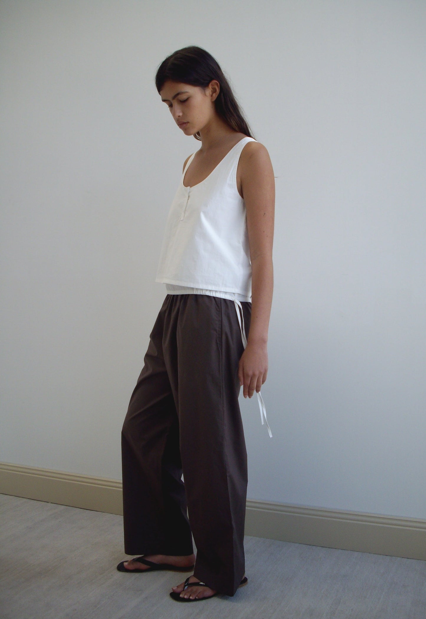 A woman with long dark hair stands against a plain wall, wearing a sleeveless white top, Ease Trouser - Chocolate by Deiji Studios in organic cotton, and black sandals. She looks down with a relaxed posture.