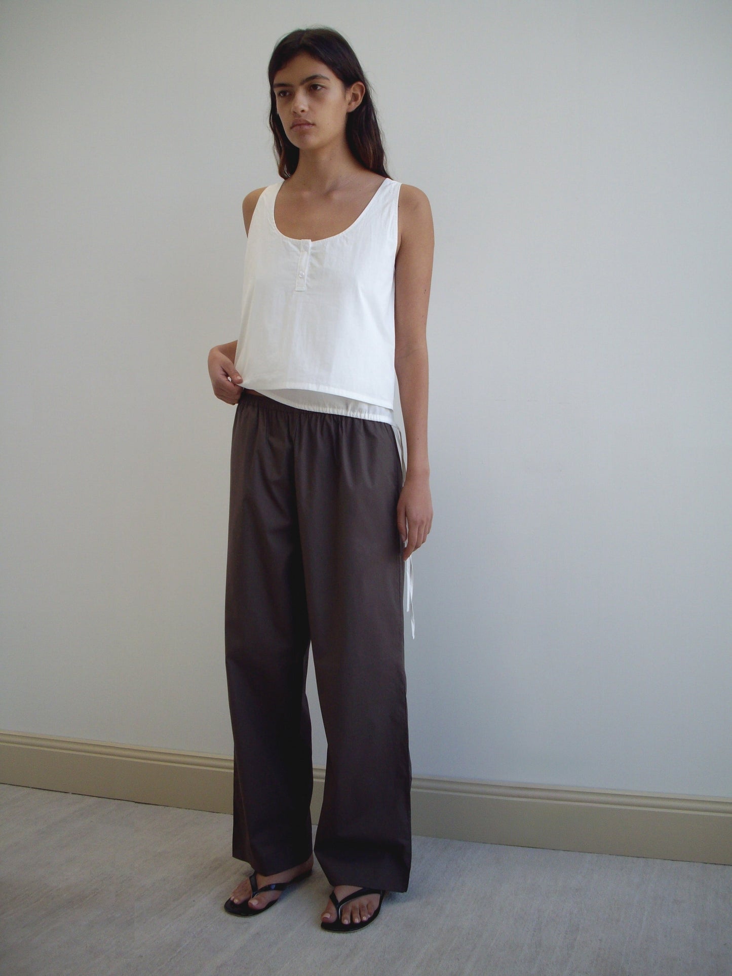 A person stands indoors against a plain wall, wearing a loose white sleeveless top, the Ease Trouser - Chocolate from Deiji Studios, and black sandals. They have long dark hair and a calm expression.