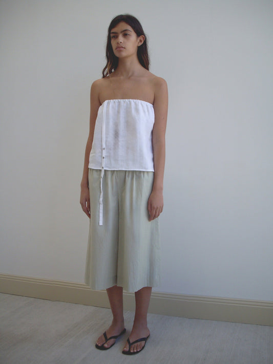 A woman stands indoors in a white strapless top and Deiji Studios Long Lounger - Pistachio Stripe pants, paired with black flip-flops. She faces forward, hands by her sides, in front of a plain wall.