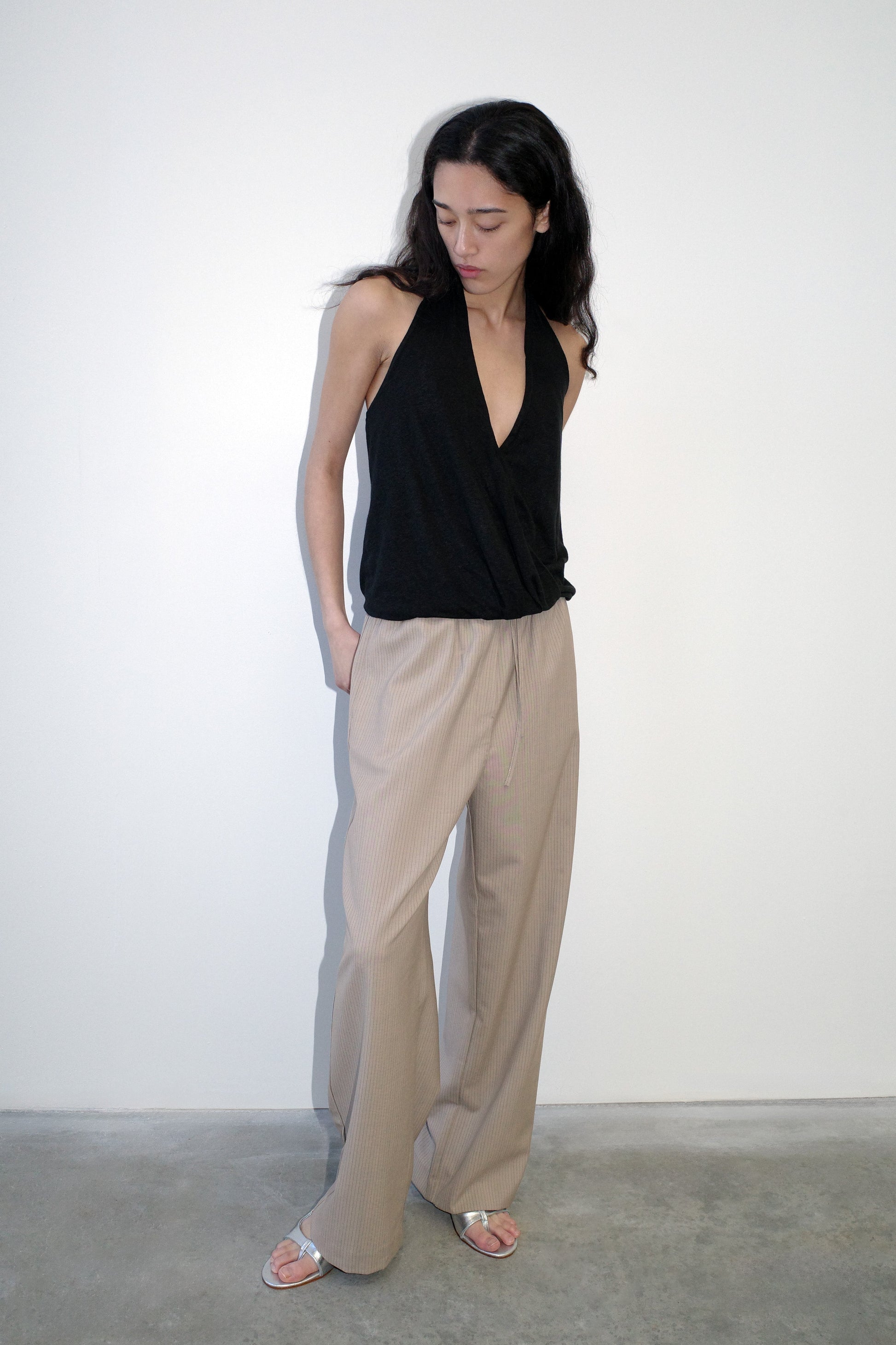 A woman with long dark hair stands against a white wall, looking down with her hands in her pockets. She wears a sleeveless black halter top, silver sandals, and Low Pocket Pure Wool Pant - Dove Stripe in a relaxed fit.