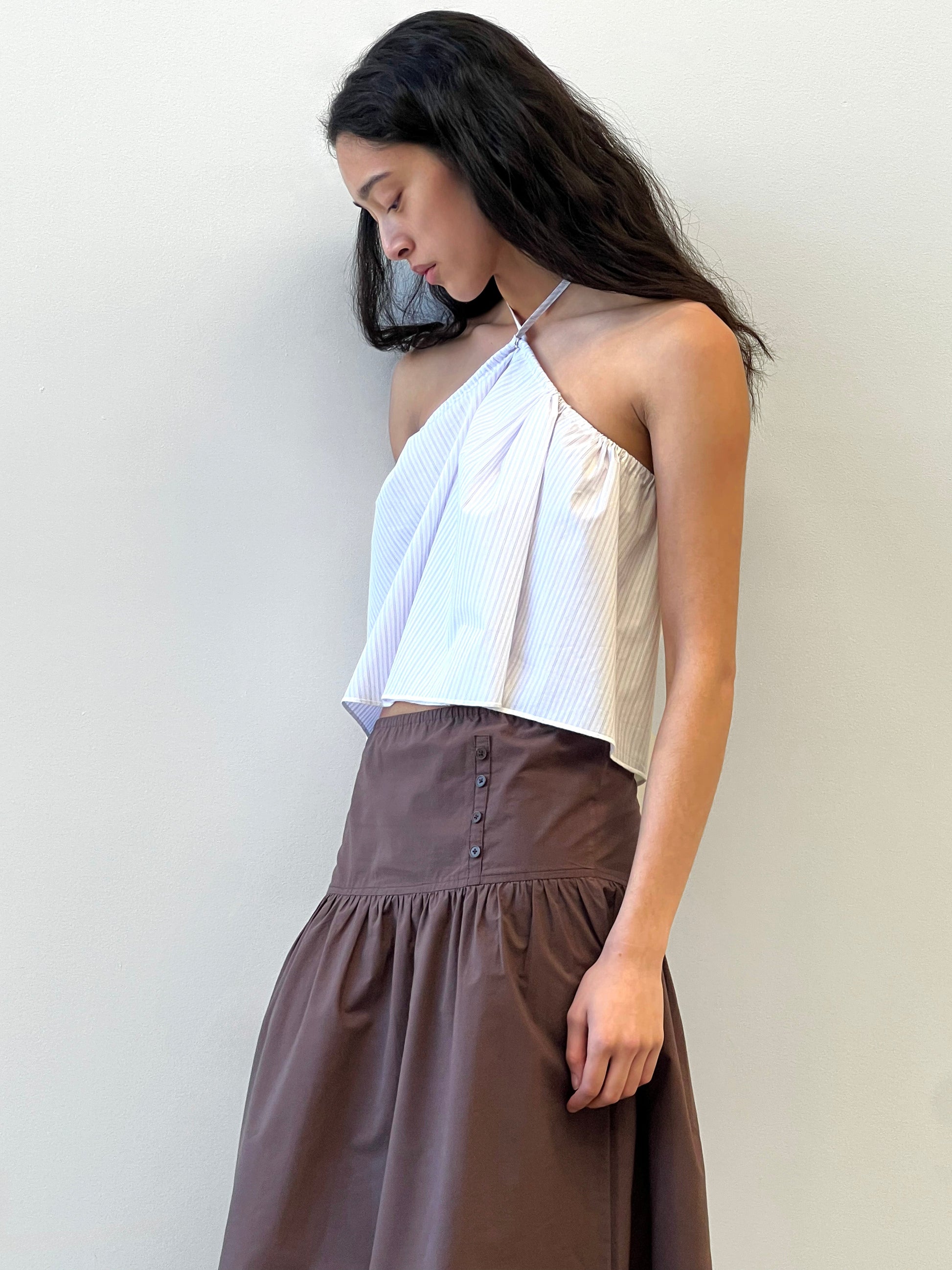 A woman with long dark hair wears a sleeveless, light halter top and the Button Skirt - Chocolate in organic cotton from Deiji Studios, standing against a plain background and looking downward.