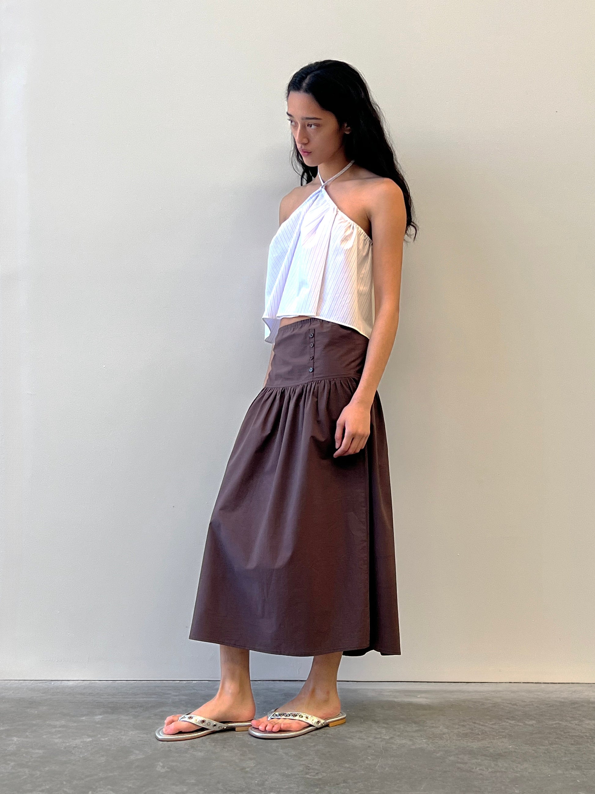 A woman with long dark hair and a neutral expression stands indoors against a plain wall, wearing a white asymmetrical halter top, the Button Skirt - Chocolate from Deiji Studios in organic cotton, and flip-flops.