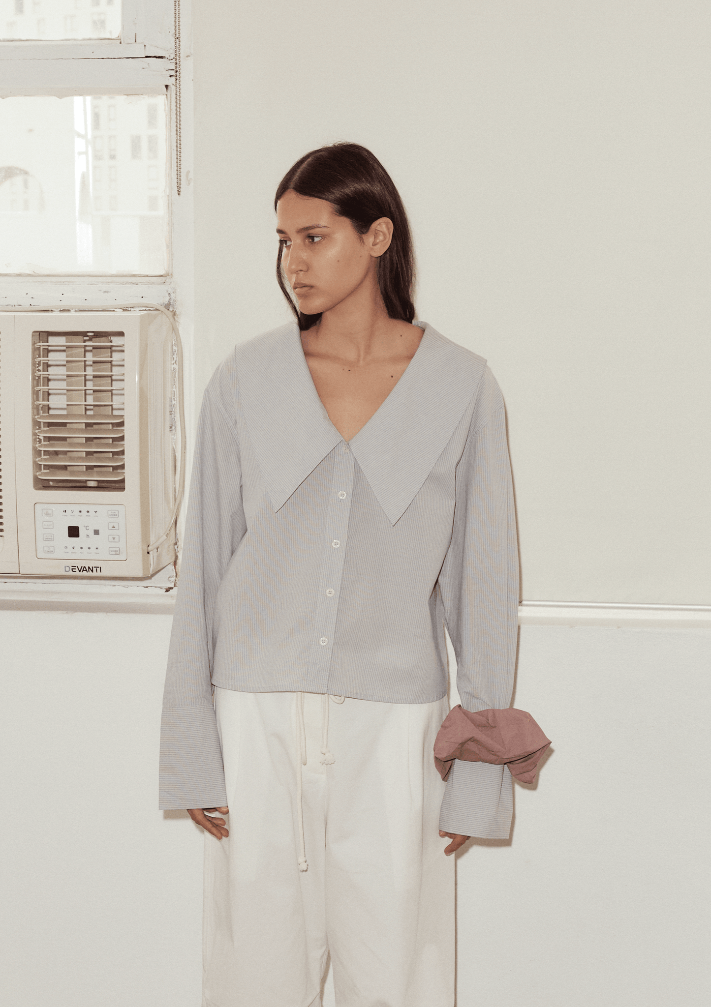 Wearing an Oversized Collared Shirt in light blue, paired with loose white pants and a mauve fabric scrunchie on the sleeve, a person with straight brown hair stands indoors by a window with an air conditioner, sporting a neutral expression.