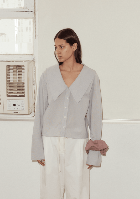 Wearing an Oversized Collared Shirt in light blue, paired with loose white pants and a mauve fabric scrunchie on the sleeve, a person with straight brown hair stands indoors by a window with an air conditioner, sporting a neutral expression.