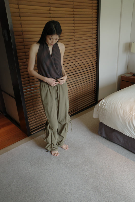 A woman with long dark hair stands barefoot in front of wooden blinds in a bedroom, wearing the Drawcord Pocket Pant - Khaki by Deiji Studios, khaki wide-leg trousers, with a brown striped halter top.