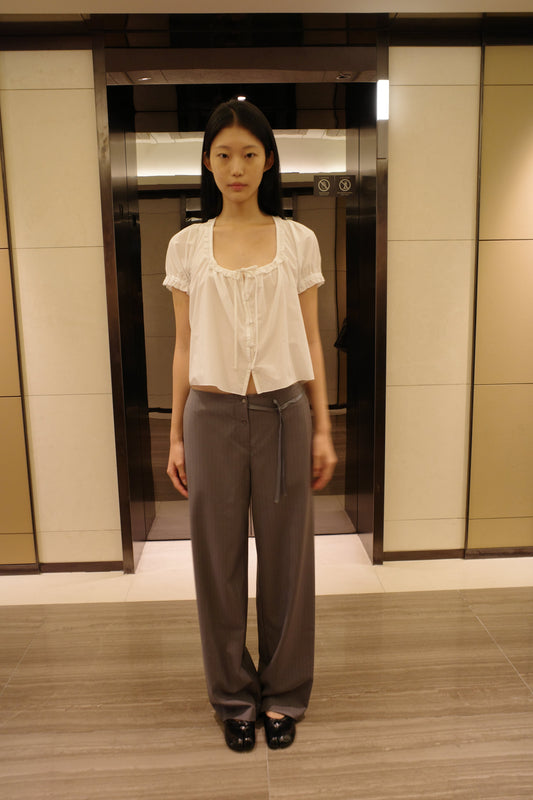 A woman with long dark hair stands in front of a lift wearing the Flounced Top - White by Deiji Studios, a white top with a frilled scoop neckline, front tie and puff sleeves, with grey pinstripe trousers and black shoes.