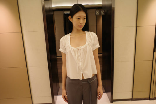 A woman with long dark hair stands in front of a lift wearing the Flounced Top - White by Deiji Studios, a white top with a frilled scoop neckline and puff sleeves, with grey pinstripe trousers.