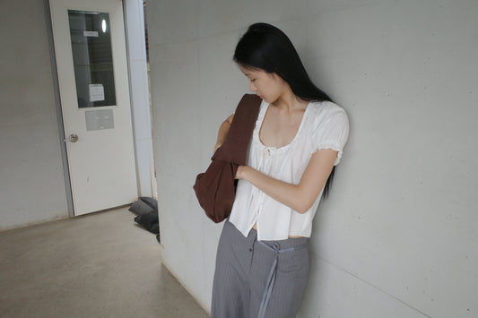 A woman with long dark hair leans against a wall in a corridor wearing the Flounced Top - White by Deiji Studios, a white top with a frilled scoop neckline and puff sleeves, with grey trousers and a brown shoulder bag.