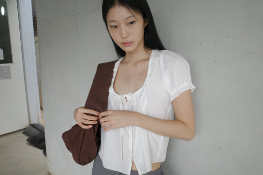 A woman with long dark hair stands in a corridor looking down, wearing the Flounced Top - White by Deiji Studios, a white top with a frilled scoop neckline, front tie and puff sleeves, with a brown shoulder bag.
