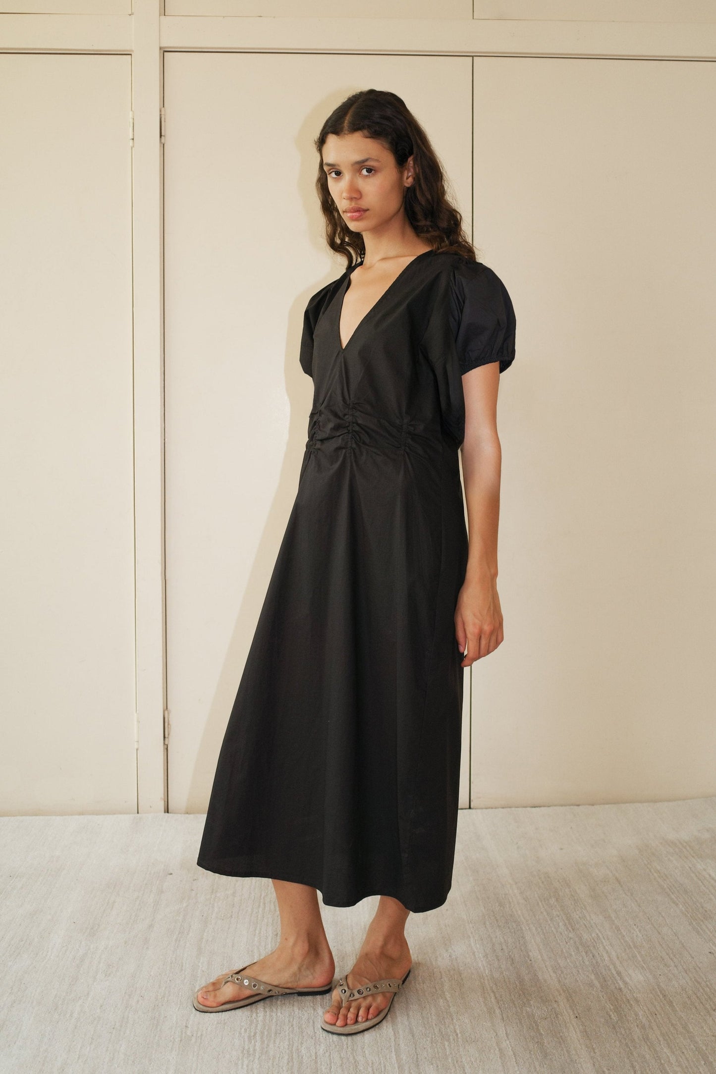 A woman with long, wavy hair wears the Pinched Dress - Black, a short-sleeved organic cotton V-neck midi dress, paired with beige flat sandals. She stands indoors before cream paneled doors.