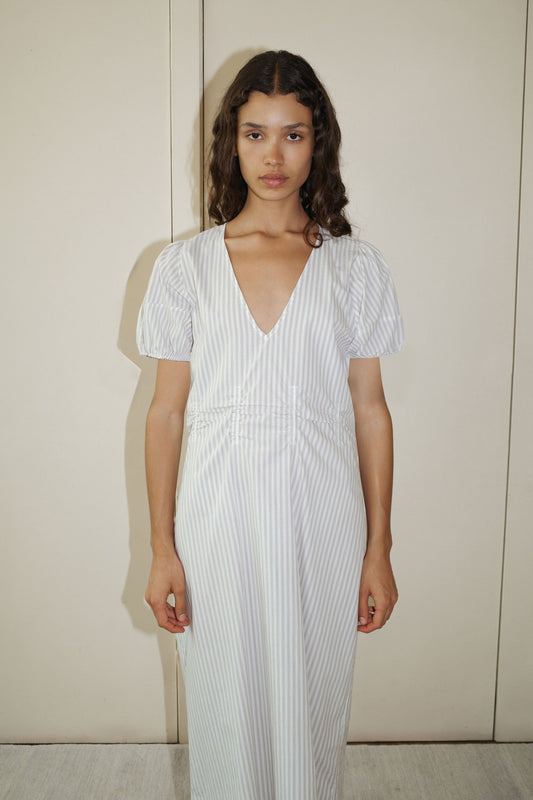 A woman with long, wavy brown hair wears the Pinched Dress - Lake Stripe, a light gray and white striped organic cotton poplin dress with short puff sleeves and a deep V-neckline, standing against a plain beige wall.