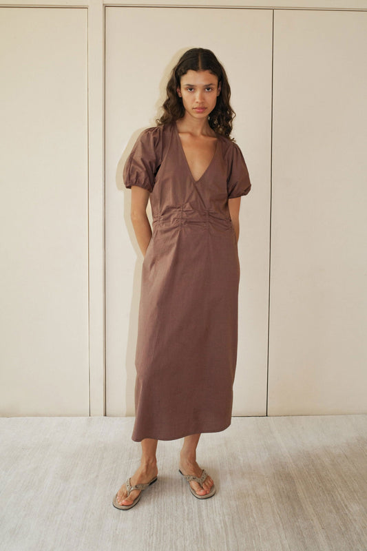 A woman with long curly hair wears the Pinched Dress—a brown, short-sleeved, V-neck midi—paired with beige sandals, standing indoors against a beige wall and light floor.