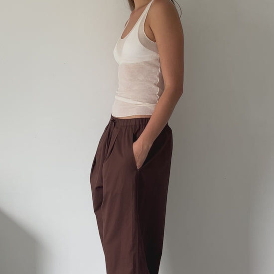 female model wears soft cotton trouser featuring a drawcord hem and waist with deep side pockets designed to sit mid-low waist for a relaxed look and made from 100% GOTS certified organic cotton. Paired with a knit tank in off white sheer fine knit tank with a low curved neckline and racer style back in a form fit, features an asymmetric baby lock exposed hem detail and finishes, made from a responsible blend of 65% ecovero viscose and 35% recycled polyester with OEKO-TEX 100 certification. 