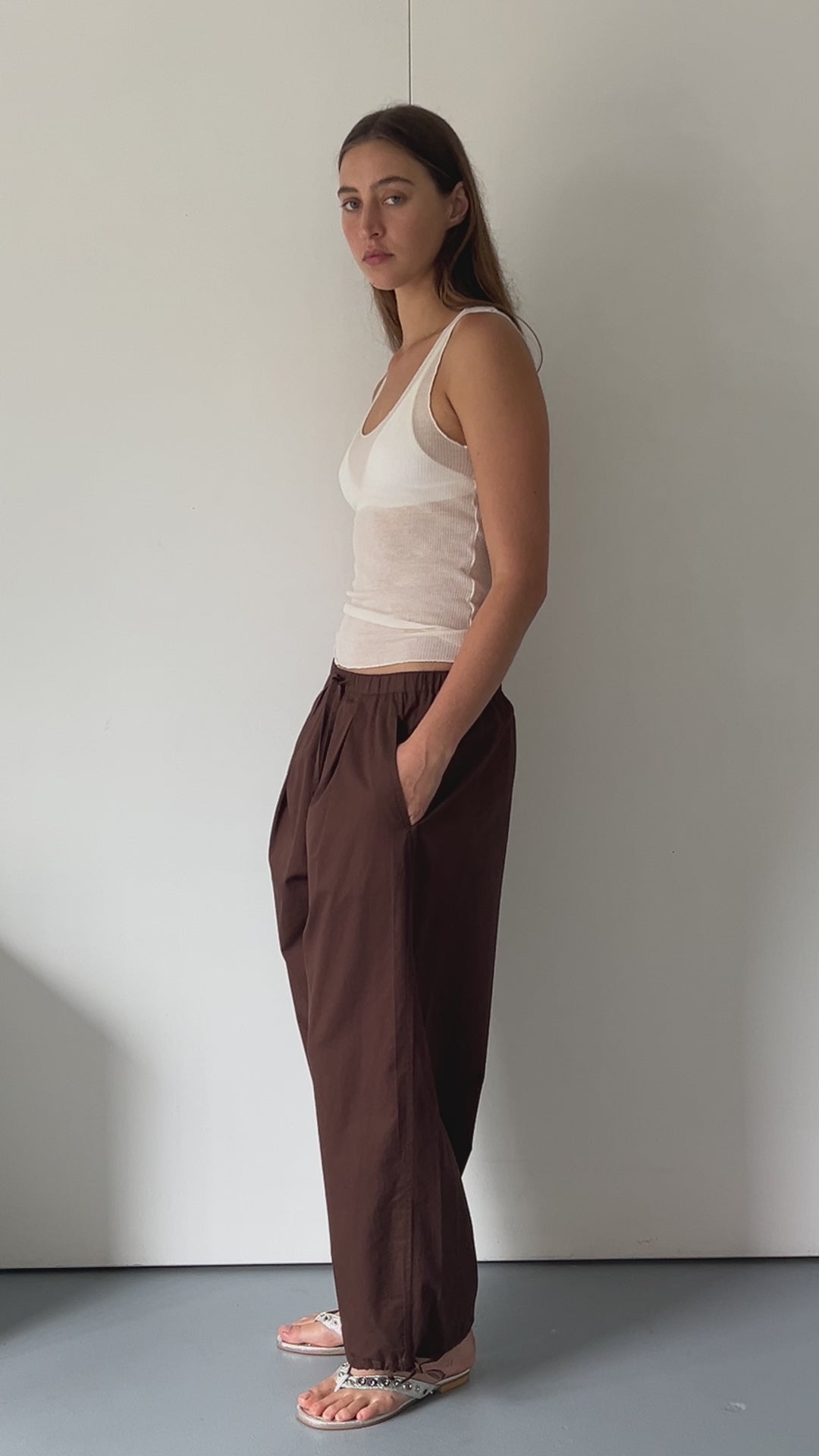 female model wears soft cotton trouser featuring a drawcord hem and waist with deep side pockets designed to sit mid-low waist for a relaxed look and made from 100% GOTS certified organic cotton. Paired with a knit tank in off white sheer fine knit tank with a low curved neckline and racer style back in a form fit, features an asymmetric baby lock exposed hem detail and finishes, made from a responsible blend of 65% ecovero viscose and 35% recycled polyester with OEKO-TEX 100 certification. 