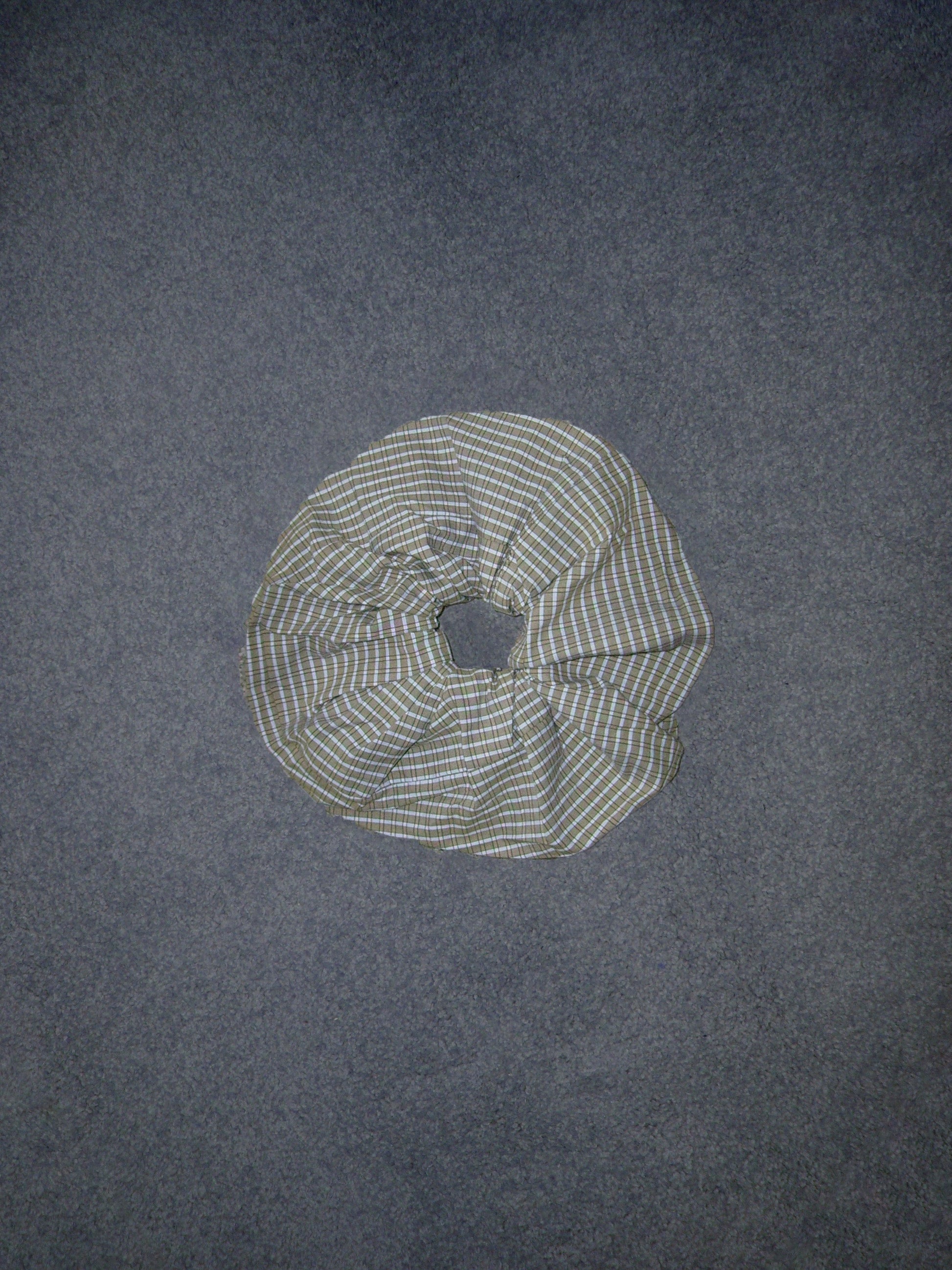 A Deiji Studios Scrunchie featuring green, blue, and white plaid fabric rests on a flat, dark gray surface.