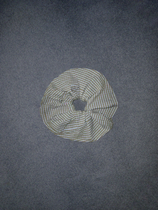 A Deiji Studios Scrunchie featuring green, blue, and white plaid fabric rests on a flat, dark gray surface.