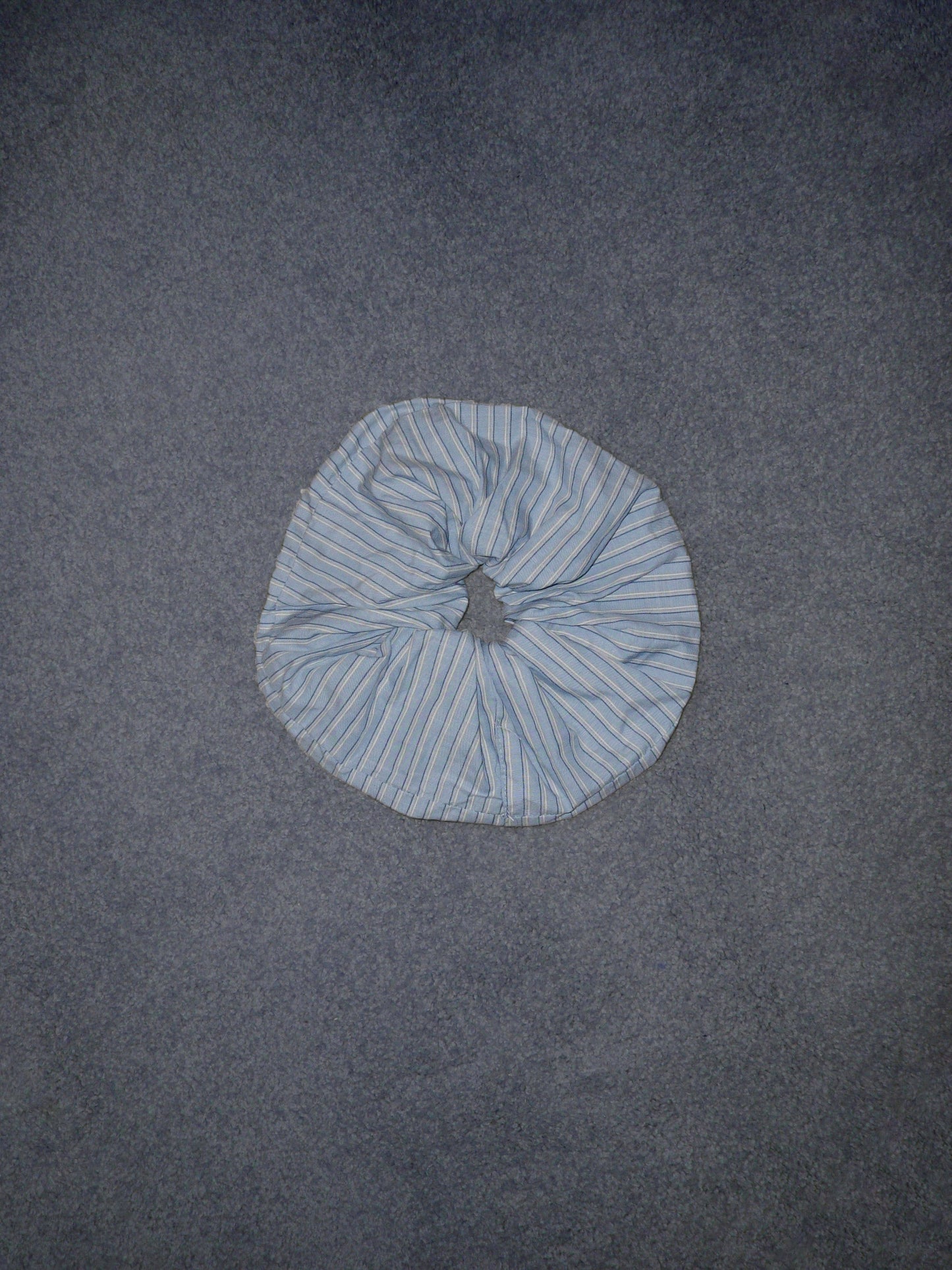 A Scrunchie in light blue and white stripes rests on a plain, textured gray surface.