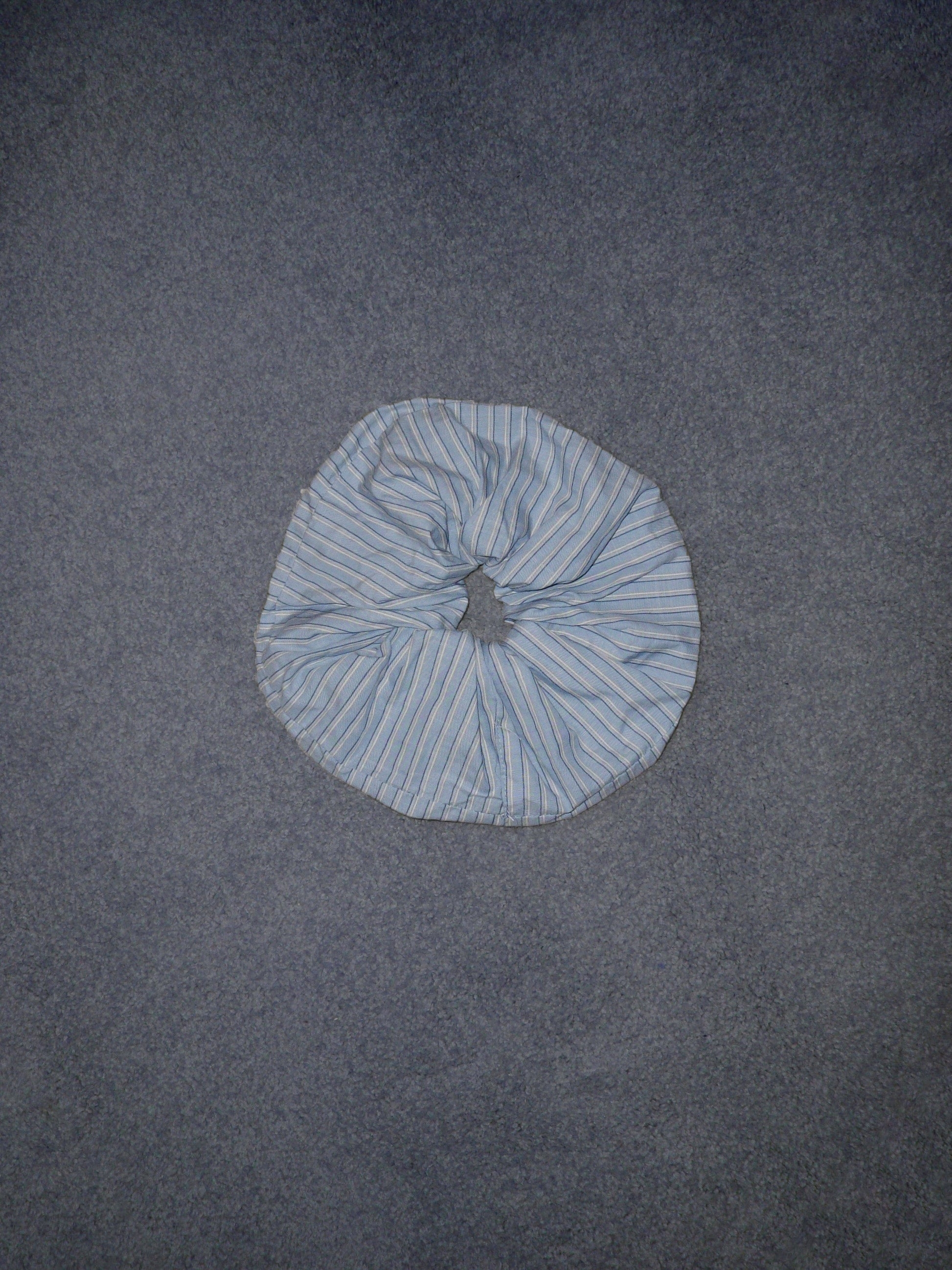 A Scrunchie in light blue and white stripes rests on a plain, textured gray surface.