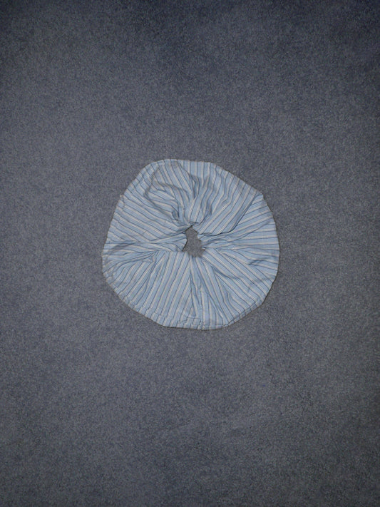 A Scrunchie in light blue and white stripes rests on a plain, textured gray surface.