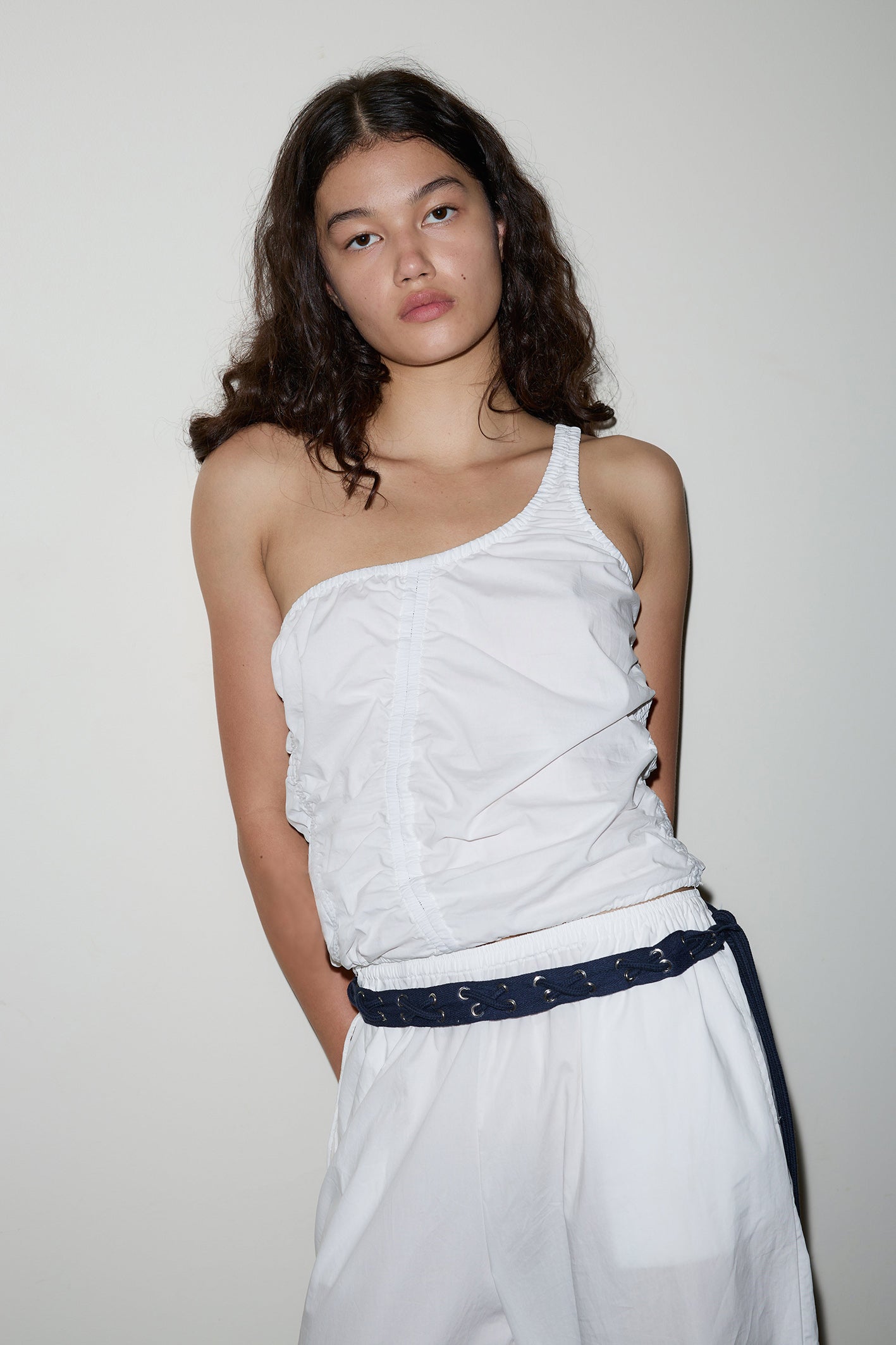 A woman with long, wavy dark hair stands against a white wall, wearing the Scrunchie Top—an elegant white one-shoulder piece—paired with loose white pants and a dark belt. Her hands are behind her back and she has a neutral expression.