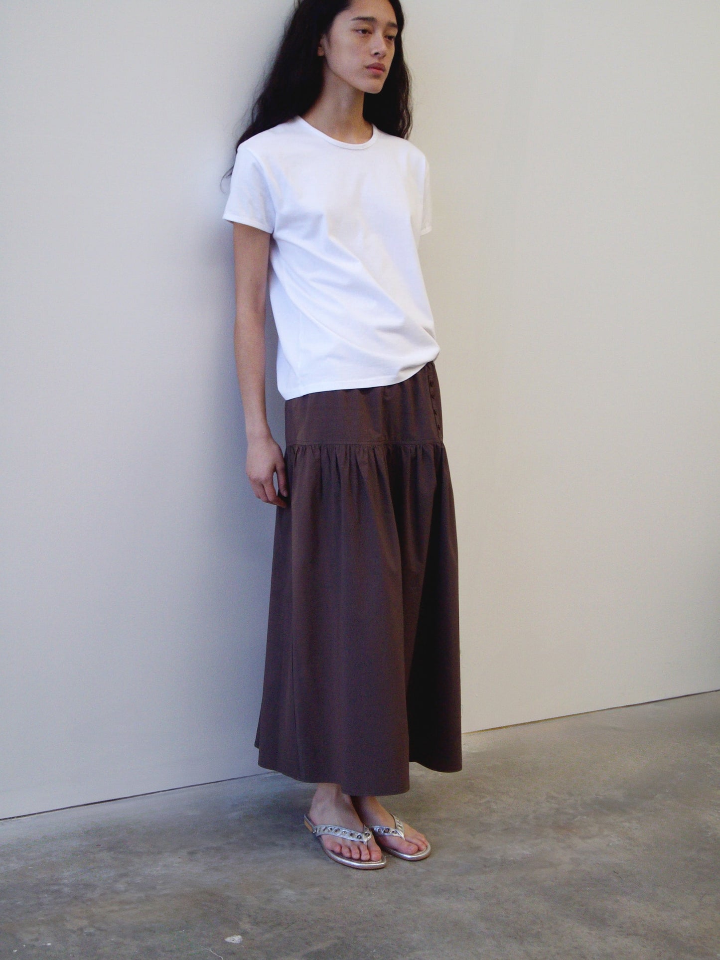 A person with long dark hair wears a white t-shirt, the Deiji Studios Button Skirt in Chocolate, and sandals while standing against a plain white wall on a concrete floor.