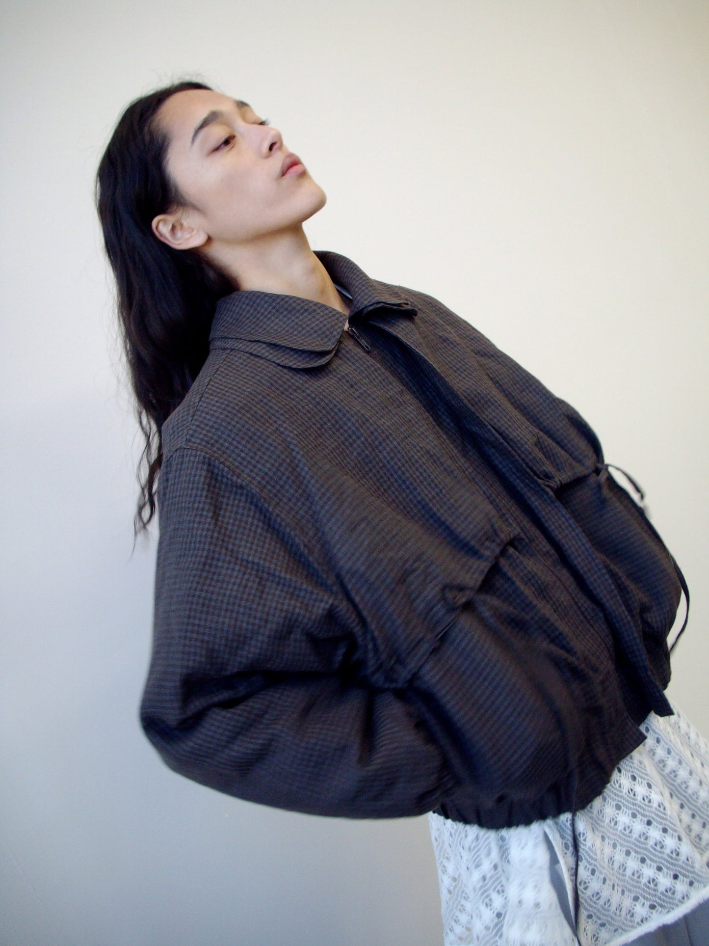 A person with long dark hair, slightly leaning back, wears the Double Layered Jacket - Slate Check from Deiji Studios and patterned light-colored pants against a plain light background.