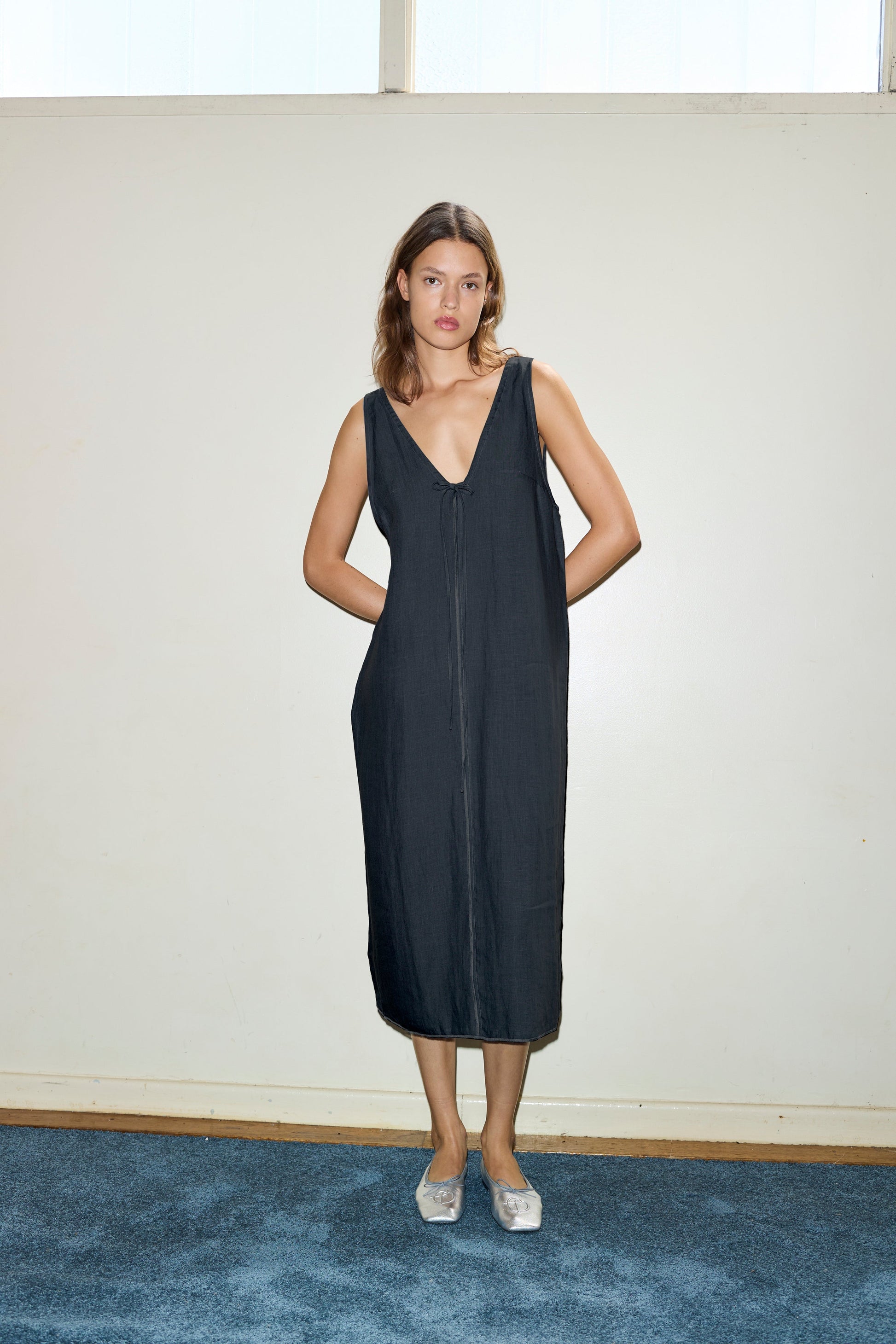 A woman stands indoors against an off-white wall, wearing the sleeveless Tie Slip Dress in black with a deep V-neck. She has her hands in her pockets and is paired with silver slip-on shoes on blue carpet.