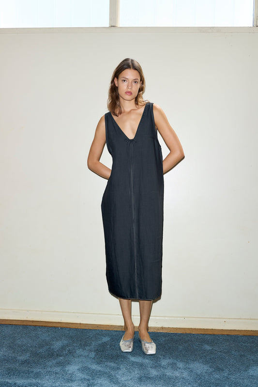A woman stands indoors against an off-white wall, wearing the sleeveless Tie Slip Dress in black with a deep V-neck. She has her hands in her pockets and is paired with silver slip-on shoes on blue carpet.