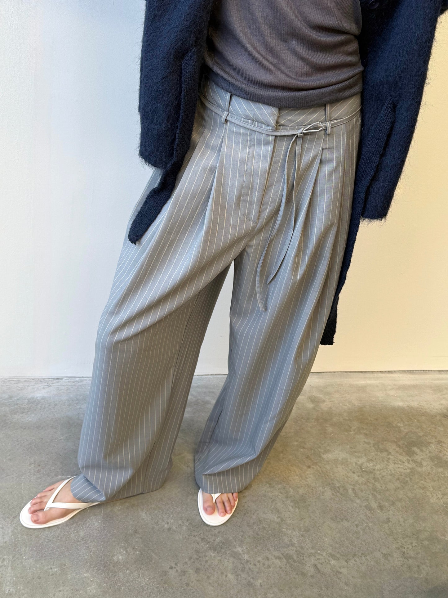 A person wearing the Tri Pleat Pure Wool Pant - Steele Stripe with soft front pleats, a dark top, navy cardigan, and white flip-flops stands on a concrete floor against a light-colored wall.